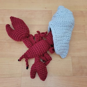 May include: A crocheted lobster toy with a red body and black eyes. The lobster is wearing a gray crocheted hat.