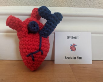 May include: A crocheted red heart with blue veins and a blue crocheted aorta. The heart is sitting on a wooden surface next to a white card with the text "My Heart Beats for You".