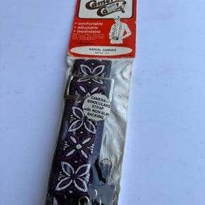 May include: A Camera Catch brand casual carrier camera and binoculars strap, style 711, in original plastic packaging. The strap features a purple and black geometric floral pattern with a silver metal buckle and non-slip backing.