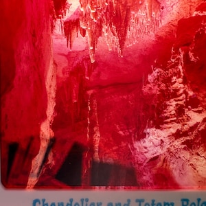 Può includere: Una fotografia dai toni rosso vibrante di un interno di grotta con stalattiti pendenti e una formazione centrale di stalagmiti. L'immagine include il testo blu stampato Chandelier and Totem Pole in basso, che cattura le caratteristiche geologiche.
