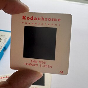 May include: A square Kodachrome transparency slide with a white cardboard frame. The text Kodachrome and TRANSPARENCY are printed in red at the top. The text THIS SIDE TOWARD SCREEN is printed in red at the bottom, with the letters AL in the corner.