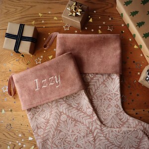 Op de afbeelding: Twee kerstsokken met een bloemenpatroon en een roze manchet, één geborduurd met de naam "Izzy". De sokken liggen op een houten oppervlak met ingepakte cadeaus en gouden en zilveren ster- en boomconfetti.