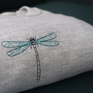 Può includere: Una felpa grigia con un ricamo a forma di libellula. La libellula è nera con ali turchesi.