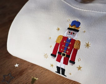 May include: Cream-colored sweatshirt featuring an embroidered Nutcracker soldier design. The soldier is dressed in a red jacket, blue hat, and black boots, with gold accents. Gold star embroidery surrounds the Nutcracker. A festive, holiday-themed garment.