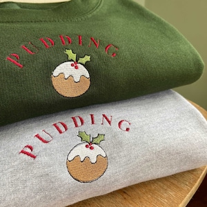 Könnte beinhalten: Zwei Weihnachtspullover, einer grün und einer grau, mit einem gestickten Weihnachtspudding und dem Wort "PUDDING" in Rot.