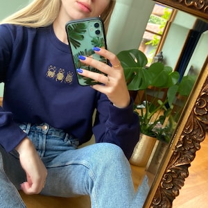 Könnte beinhalten: Eine Frau trägt einen marineblauen Pullover mit einem goldenen Stickmuster aus drei Marienkäfern. Sie trägt eine blaue Jeans und hat blauen Nagellack. Sie hält eine grüne und goldene Handyhülle mit einem tropischen Blattdesign.