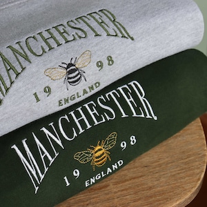 May include: Two sweatshirts stacked on top of each other. The top sweatshirt is grey with green embroidery that says "MANCHESTER 1998 ENGLAND" with a bee graphic. The bottom sweatshirt is green with white embroidery that says "MANCHESTER 1998 ENGLAND" with a bee graphic.