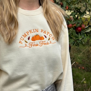 Könnte beinhalten: Cremefarbenes Sweatshirt mit gesticktem Text und Grafiken. Das Design zeigt die Worte "PUMPKIN PATCH Farm Fresh" in orangefarbenem Faden, zusammen mit orangefarbenen Kürbissen und Herbstblättern. Das Sweatshirt ist langärmlig und hat einen Rundhalsausschnitt.