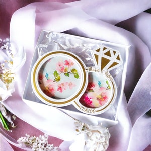 Könnte beinhalten: Weiße Ringbox mit einem goldenen Ringdesign. Der Ring ist mit floralen Mustern und den Worten "Maria & Anton" und "21.08.2019" verziert.