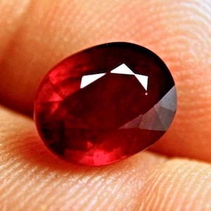 May include: A deep red, oval-cut gemstone with a faceted surface that reflects light. The stone displays a rich, saturated crimson hue and is polished to a high shine, showcasing internal clarity and precise geometric cuts across its top surface.