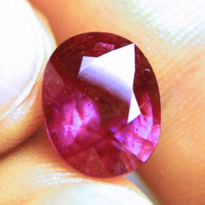 May include: A faceted oval-shaped gemstone with a deep magenta and red colour profile. The stone features a polished surface with multiple geometric cuts that reflect light, creating bright white highlights across its translucent, vibrant surface.