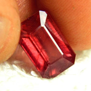 May include: A rectangular, emerald-cut gemstone with a deep, vibrant red color. The stone features multiple facets that reflect light, creating bright highlights and dark shadows across its surface, showcasing a clear, transparent crystalline structure.