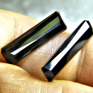 Puede incluir: Dos cristales de piedras preciosas negras facetadas. Los cristales tienen forma rectangular con lados angulados y superficies pulidas, que reflejan la luz. Las piedras preciosas se muestran sobre un fondo neutro.