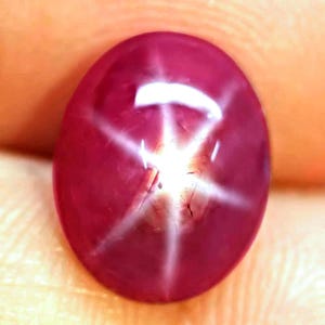 May include: A deep magenta star ruby gemstone with a smooth, polished cabochon cut. The stone features a distinct six-rayed white star effect, known as asterism, centered on its surface, reflecting light with a bright, sharp, and luminous pattern.