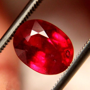 May include: A faceted, oval-shaped ruby gemstone held by tweezers. The ruby is a deep, vibrant red color with internal reflections. The tweezers are silver-colored and have a serrated edge.