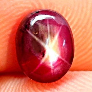 Puede incluir: Una gema de rubí pulida, de forma ovalada. La piedra de color rojo intenso presenta un efecto de estrella de seis rayos, con líneas blancas y rosa claro que irradian desde el centro. La gema tiene una superficie brillante.