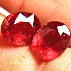 May include: Two oval-cut, deep red rubies with faceted surfaces that reflect light. The gemstones exhibit a vibrant, saturated crimson hue and a polished finish, showcasing internal clarity and distinct geometric facets across their rounded shapes.