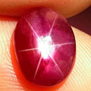 May include: A deep magenta-colored star ruby gemstone with a smooth, polished cabochon cut. The stone displays a distinct six-rayed star effect, known as asterism, which radiates from a bright central point across the curved surface of the gem.
