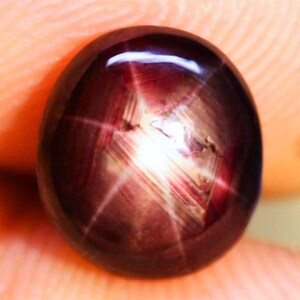 May include: A deep dark red cabochon gemstone featuring a prominent six-rayed star effect known as asterism. The oval-shaped stone displays a smooth, polished surface with a distinct light reflection that creates a bright white star pattern.