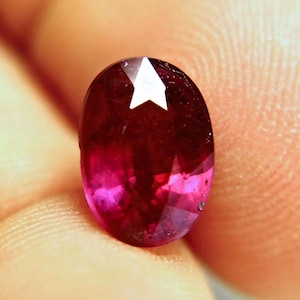 May include: An oval-cut gemstone with a deep, saturated red colour and a faceted surface. The stone exhibits internal clarity variations and a distinct white light reflection on its upper portion, showcasing its polished, translucent appearance.