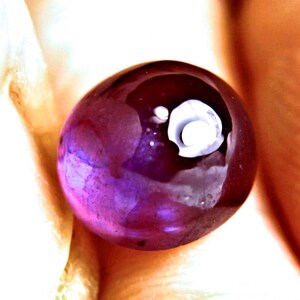 May include: A round, polished gemstone with a deep purple and magenta hue, featuring a smooth, reflective surface and a distinct, off-center white circular inclusion that appears as a small, bright, opaque spot within the translucent stone.
