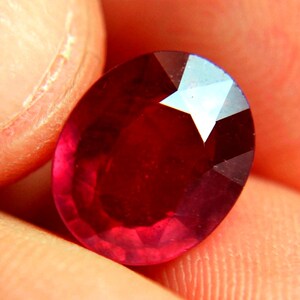 Peut inclure: Une pierre précieuse de rubis ovale rouge foncé avec une taille facetée. Le rubis est tenu dans une main, et les facettes de la pierre sont visibles.