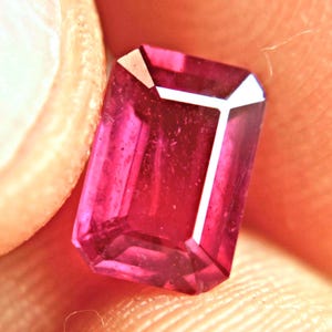 May include: A faceted, rectangular-cut, deep pink gemstone. The gem has a bright, reflective surface with white facets at the top. The gem is held between two fingers, with the skin tone providing a warm background.