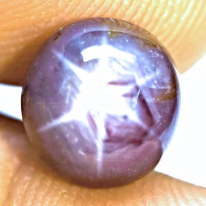 May include: A round, cabochon-cut star ruby gemstone with a deep purplish-red hue. The stone features a prominent, bright white six-rayed star effect, known as asterism, centered on its polished, smooth, and slightly translucent surface.