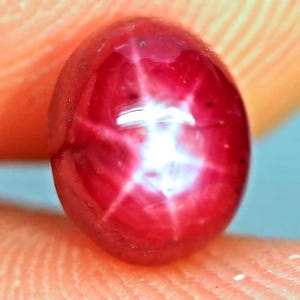 May include: A deep red, oval-shaped star ruby gemstone featuring a distinct six-rayed asterism effect. The cabochon-cut stone displays a vibrant crimson hue with a bright, centred white star reflection that shifts across the polished surface.