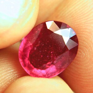 May include: A faceted oval-shaped gemstone with a deep red colour and a polished surface. The stone features multiple triangular and trapezoidal facets that reflect light, creating a bright, sparkling appearance across its translucent body.