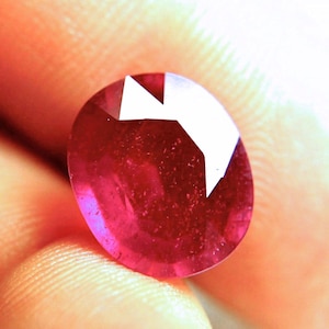 May include: A faceted oval-shaped ruby gemstone with a deep red color and a translucent appearance. The stone features multiple geometric cut surfaces that reflect light, creating bright white highlights across the top portion of the gem.
