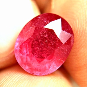 May include: A vibrant, oval-cut ruby gemstone with a deep red hue and multifaceted surface. The stone displays internal clarity and light-reflecting facets across its polished top, showcasing a rich, saturated color profile throughout the gem.