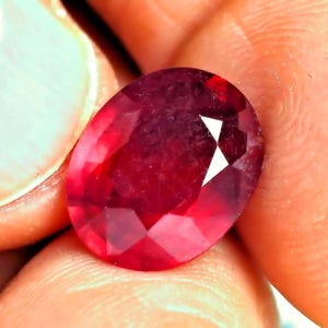 May include: A deep red, oval-cut gemstone with a faceted surface that reflects light. The stone displays a rich, saturated crimson hue throughout its transparent body, showcasing a classic oval shape with multiple geometric facets across its top.