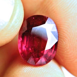 May include: A deep red, oval-cut gemstone with multiple facets that reflect light. The stone displays a rich, saturated crimson hue throughout its surface, showcasing a polished, transparent appearance with visible internal clarity and sharp edges.