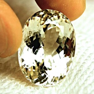 Könnte beinhalten: Ein facettierter ovaler Edelstein mit einem blassen champagnerfarbenen oder hellgelben Farbton. Der Stein weist zahlreiche geometrische Schliffe auf, die für ein hohes Maß an Brillanz und Lichtreflexion sorgen und eine klare, transparente Qualität zeigen.