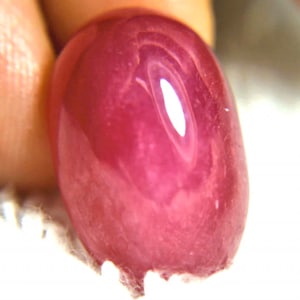 May include: A close-up of a smooth, oval-shaped, pink gemstone. The gemstone has a high polish and is reflecting light.