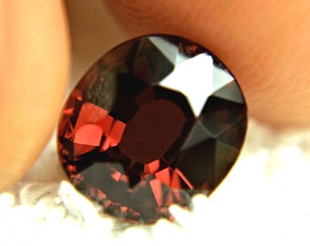 May include: A deep red, oval-cut gemstone with multiple facets. The gem's surfaces reflect light, creating a sparkling effect. The stone is held between two fingers, showing its size and colour.