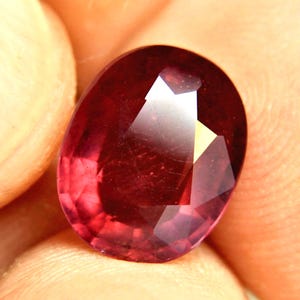 May include: A deep red, oval-cut gemstone with a faceted surface that reflects light. The stone displays a rich, saturated crimson hue with visible internal clarity and sharp geometric facets across its polished, rounded top surface.