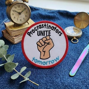 May include: A round patch with the text "Procrastinators UNITE tomorrow" encircling a raised fist. A vintage brass alarm clock, a small compass, and a pair of iridescent tweezers are also visible on a denim surface.