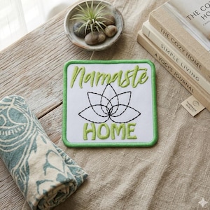 May include: Square white patch with green border and embroidered text "Namaste HOME" and a lotus flower design. A rolled-up patterned towel, a small bowl with stones, and stacked books are also visible.