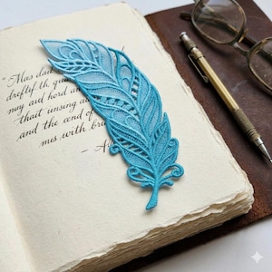 May include: A light blue, openwork feather bookmark rests on the pages of an open book. The bookmark is made of delicate lace and features intricate details. A pen and glasses are in the background. The book has handwritten text.