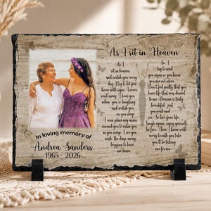 May include: A rectangular slate memorial plaque featuring a photograph of two individuals, the text As I sit in Heaven, and the name Andrea Sanders with the dates 1965-2026. The background has a wood grain pattern with butterfly illustrations.