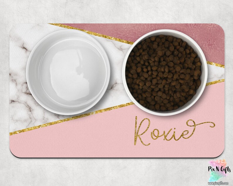 Op de afbeelding: Roze en witte marmeren voerbakset voor huisdieren met gouden accenten. De bakken zijn van wit keramiek met een roze en gouden geometrisch ontwerp. De set bevat de tekst "Roxie" in gouden glitter.