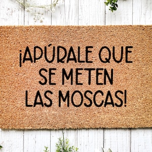 May include: A rectangular doormat made from natural coir fibre. The mat has black text that reads "¡APÚRALE QUE SE METEN LAS MOSCAS!" on a light brown background. Decorative greenery surrounds the mat.