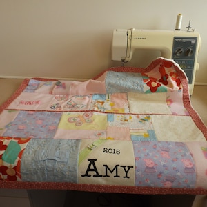 May include: A pink and white patchwork quilt with a red border. The quilt is made of various fabrics, including floral prints, butterflies, and polka dots. The quilt is personalised with the name "Amy" and the year "2015" embroidered in black thread.
