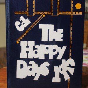 May include: A dark blue hardcover book titled The Happy Days of with white lettering and yellow graphic line accents. The cover features a stylized design and the text James Jess, Superintendent printed in small white letters at the bottom.