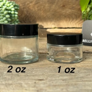 May include: Two clear glass jars with black lids. The larger jar is labelled 2 oz and the smaller jar is labelled 1 oz.