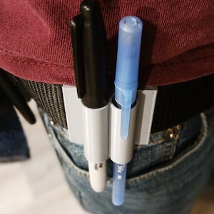 May include: A gray plastic belt clip with two pen holders. A black marker and a blue pen are inserted into the holders. The text "FINE POINT" is visible on the marker and "SHIC M" is visible on the pen.