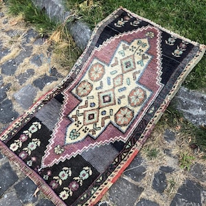 Op de afbeelding: Een rechthoekig, handgeweven tapijt met een centraal geometrisch ontwerp in cr&egrave;me, paars en teal. Het tapijt heeft een zwarte rand met bloempatronen en wordt buiten op een geplaveide ondergrond getoond.