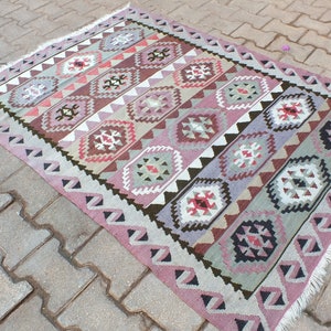 May include: A rectangular, hand-woven rug with a geometric pattern. The rug features a repeating design of diamond and rectangular shapes in shades of pink, brown, gray, and white. The edges are finished with a fringe.